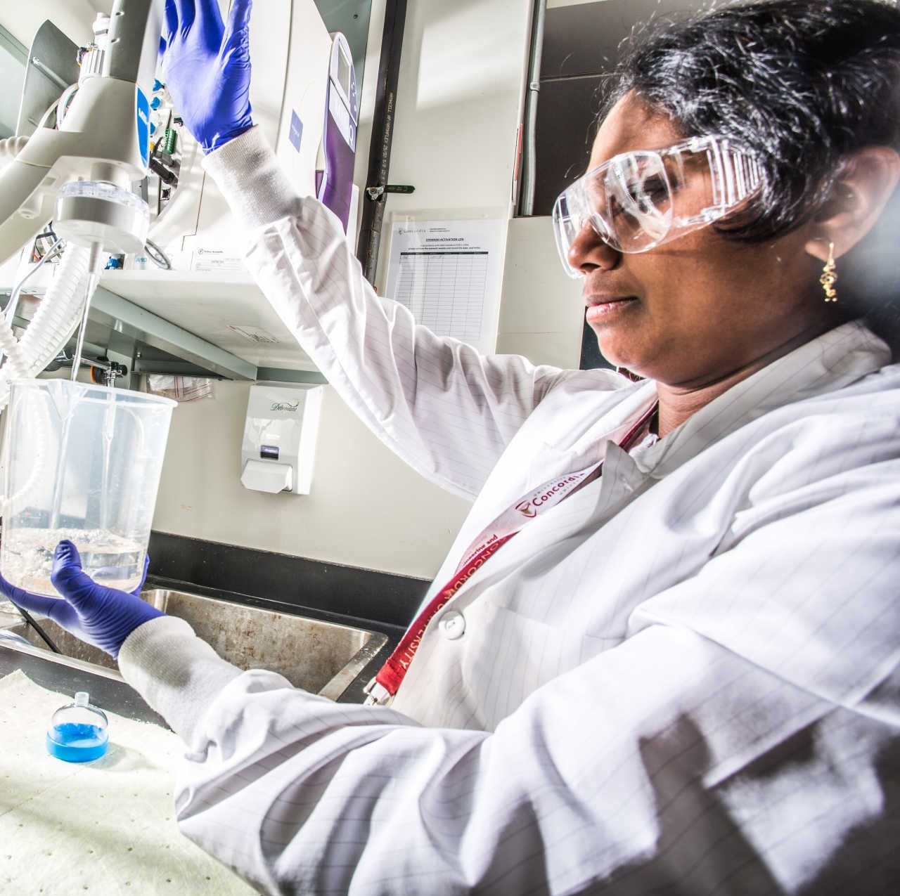 A woman in lab gear and a lab setting uses a mixer to stir a beaker with fluid in it.