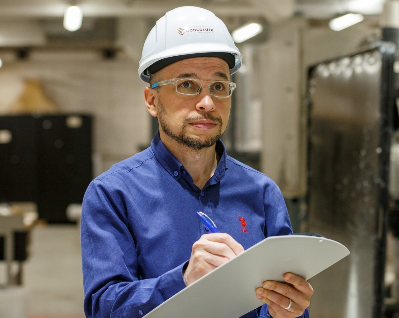 Safety Ambassador Awards A person wearing a hard hat and safety goggles and holding a clipboard moves through an industrial area where several machines and pieces of equipment are located.