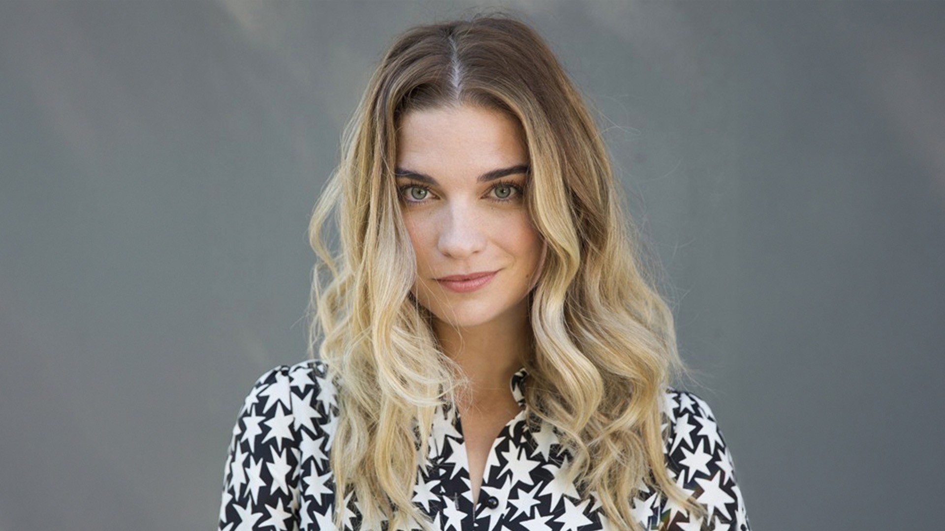 Headshot of a woman with blonde wavy hair who wearing a black and white patterned shirt as she stands in front of  grey background