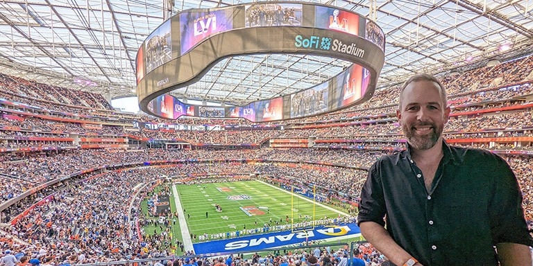 alt= "Vincent is standing in a huge stadium packed with attendees: the SoFi stadium in California. Behind him is the football field where the LA Rams are playing a game."
