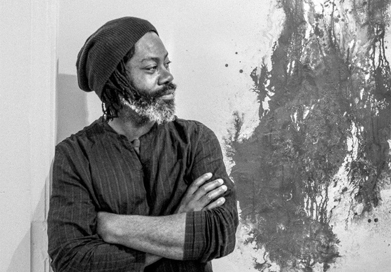 Stanley Wany, a graduate of the visual and media arts master’s program at the Université du Québec à Montréal (UQAM). Profile of a man with a beard, wearing a woollen beanie, a striped top, standing beside an abstract painted artwork.