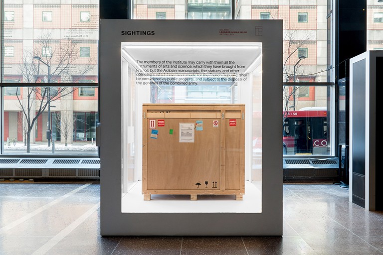 Installation view of <em>Sightings 42: approximately 760 kg of public property,</em> by Pedro Barbáchano, 2024. | Courtesy of the Leonard & Bina Ellen Art Gallery. Photo: Jean-Michael Seminar An exhibition art piece: A large square glass cube with a wooden crate inside it, in a public interior space.